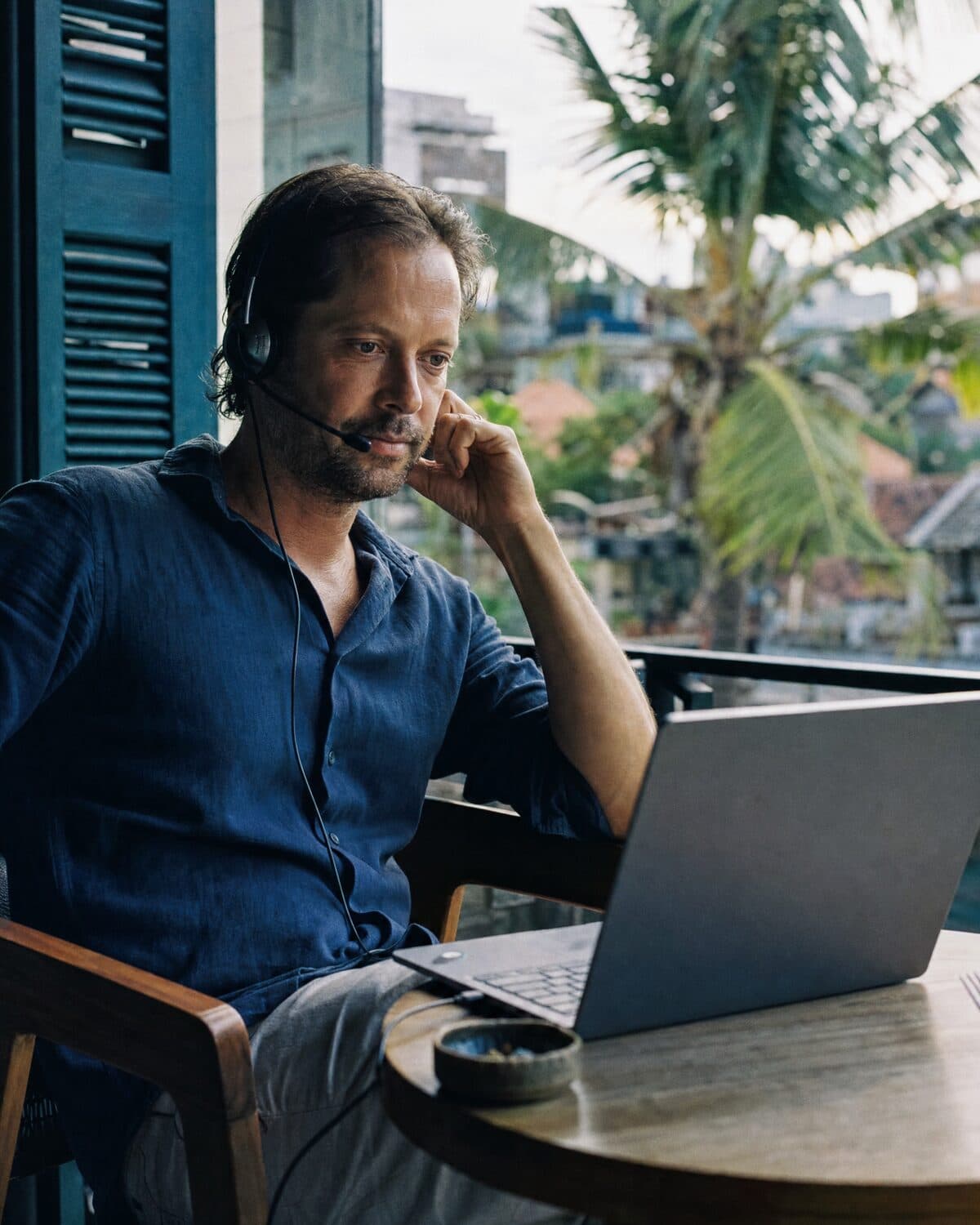A man with a headset on a call from his laptop on a balcony in a tropical city