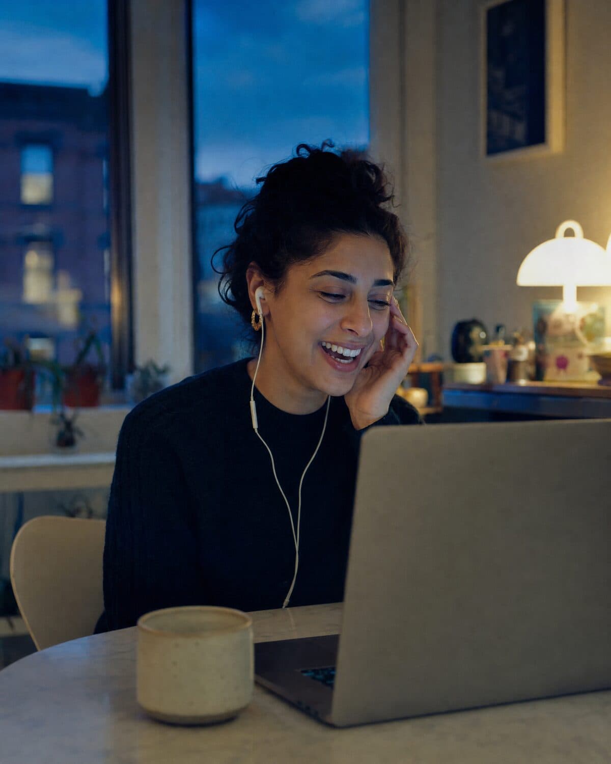 A woman laughing on a call from her laptop at the kitchen table in the evening